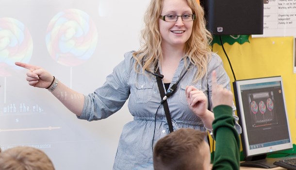 Young, smiling, female teacher in front of whiteboard in classroom. Young child with hand up.