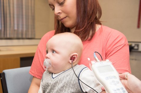 A baby sits on his mum's lap with headphones in his ears while undergoing the newborn hearing screening.