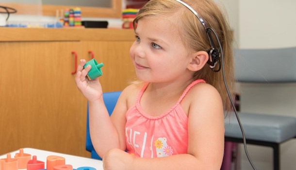 A young girl plays with blocks at an audiologist's clinic while wearing a monitoring headband.  