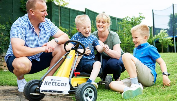 A boy rides in a small go-kart with his mum, dad and brother sitting in the grass beside him. 