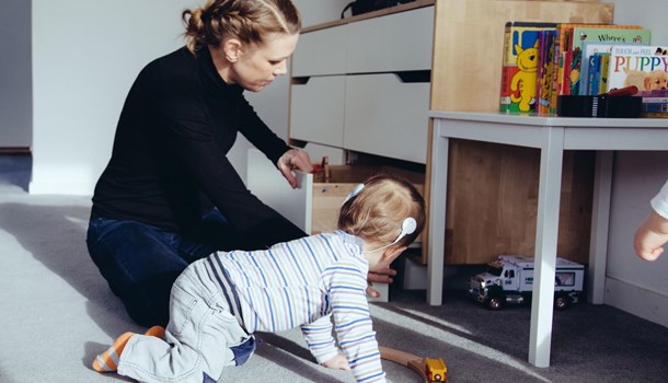 A baby wearing cochlear implants crawls next to its mum who is opening a dresser drawer.