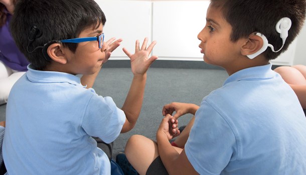 Two boys wearing cochlear implants sit next to each other, one making a motion with his hands.
