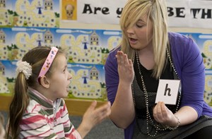 A teacher holds an alphabet card and works on phonics with a girl. 