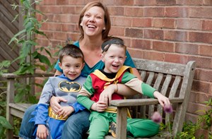 A mum holds her two sons who are wearing superhero costumes on an outdoor bench.