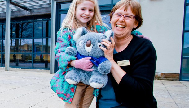 A smiling woman kneels next to a girl holding a stuffed koala bear.