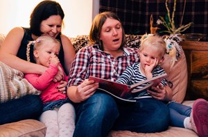 Mums sat on a sofa reading a picture book to their young twin daughters.