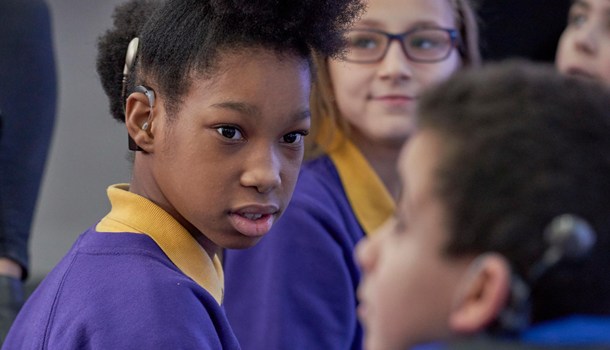 A girl in school uniform, wearing a cochlear implant, looks towards the camera at a boy also wearing a cochlear implant.