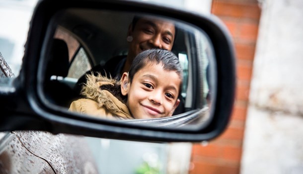 The reflection of a boy with hearing aids and his dad in a car wing mirror.