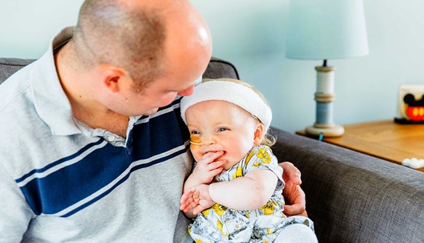 A man wearing a grey shirt with a blue stripe across it sat on a sofa holding a smiling baby 