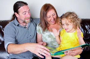 Two parents read a picture book to their toddler on the sofa. 