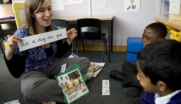 A teacher sits cross-legged in front of two pupils holding a phonics card up.