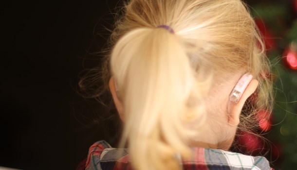 Child with blonde hair facing away from the camera, their hearing aid can be seen on their right ear.