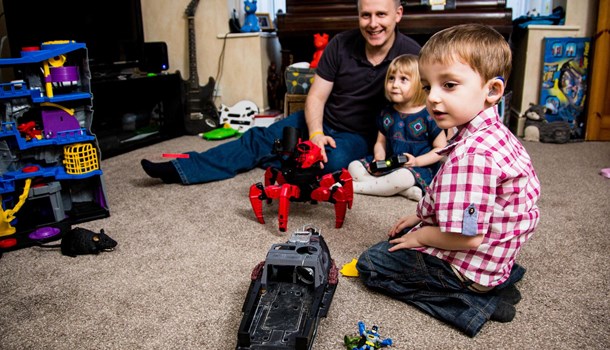 A deaf boy with blue hearing aids sat playing with toys on the floor with his dad and sister.