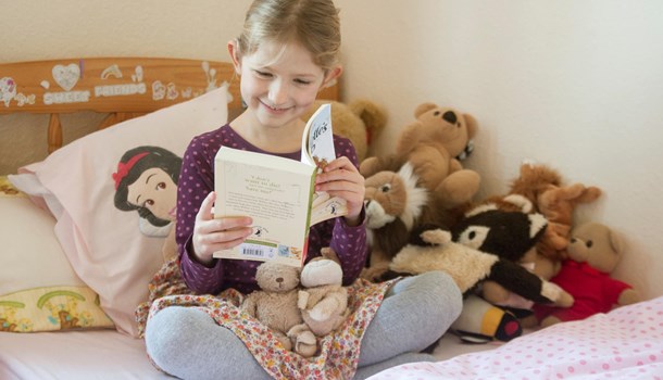Isla wearing her hearing aids sits on her bed reading a book.