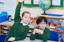 Two pupils in school uniform sit next to each other, one raising her hands.