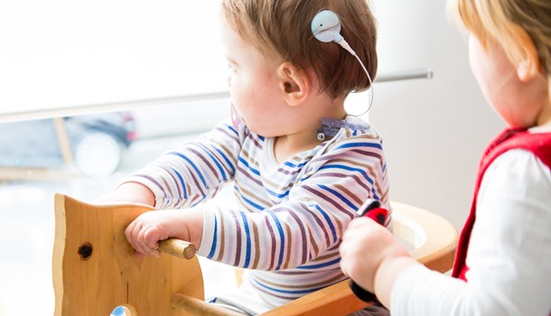 A baby wearing a hearing implant sits on a rocking horse. 