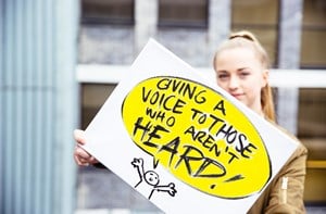 Deaf young person holding up a banner that says, 'Giving a voice to those who aren't heard!'
