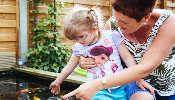 A young girl wearing a cochlear implant sits on her mom's lap while dipping her hand in a koi pond.