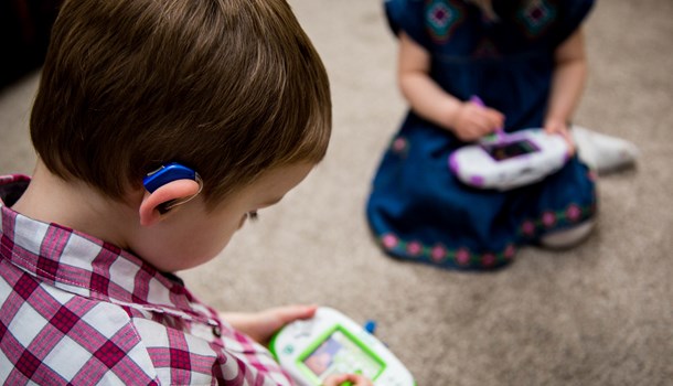 A young boy wearing a hearing aid plays with an electronic entertainment device. 