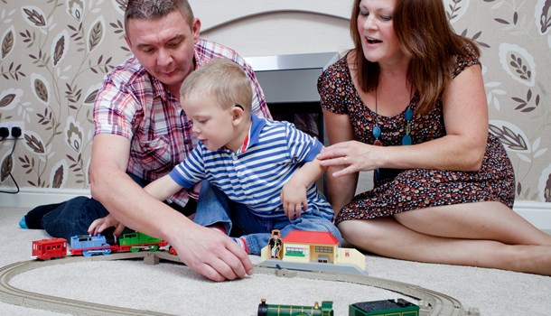 A mom and dad and their young son play with a toy train set together - the son is wearing hearing aids. 