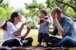 A mum and dad signing to their deaf toddler who has hearing aids in the park.