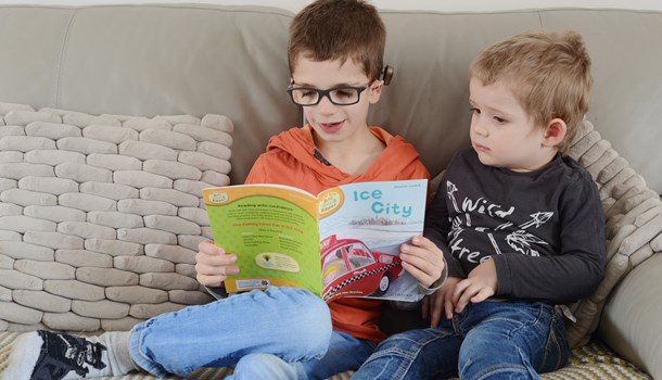 A boy wearing a hearing implant reads a picture book on the sofa next to his little brother.