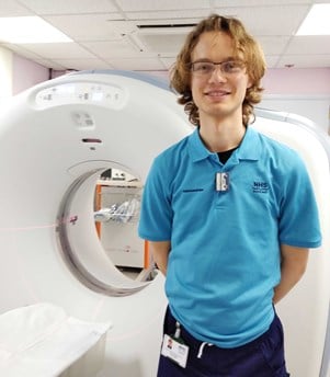 Man wearing glasses and a light blue polo top standing in front of a hospital X-ray machine.