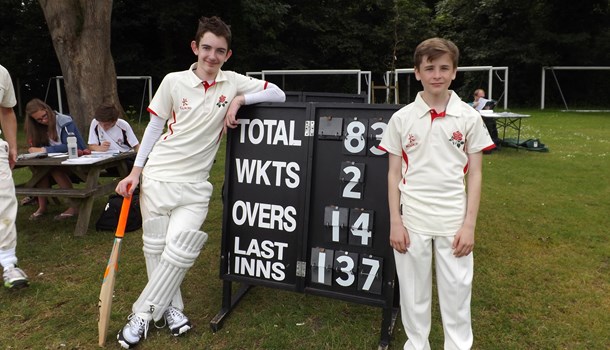 Two boys wearing cricket kit while standing next to a score board