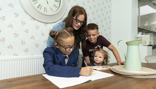Lily doing homework with her mum and siblings standing nearby