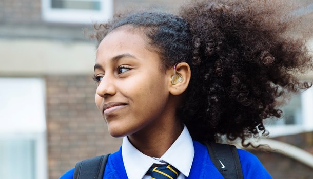 A girl with a hearing aid wears a blue school uniform and smiles. 