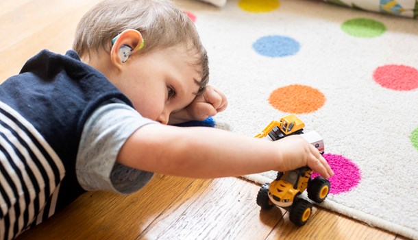 A toddler with a hearing aid plays with a toy truck on the floor. 