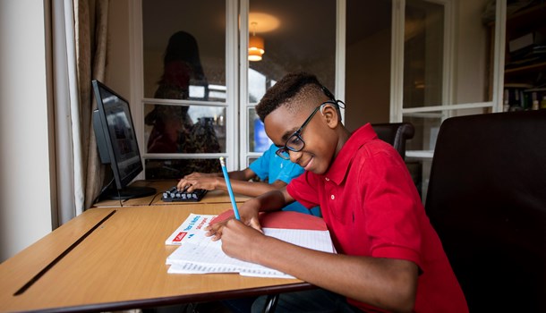 A smiling boy with cochlear implants and glasses working on his homework at a desk.