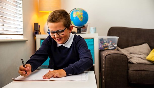 A boy with cochlear implants and glasses working on his homework at a desk with a globe in the background.