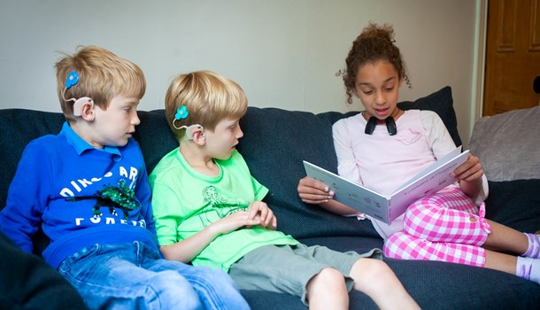 A girl reads a book on the sofa next to two boys wearing cochlear implants. 