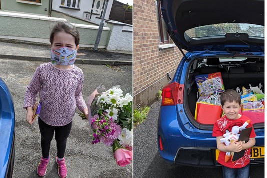 Little girl on left wearing a facemask holding flowers, boy on right holding treats standing in front of a car