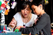 A woman with a loop around her neck looks at an art activity table with a girl wearing a hearing aid.