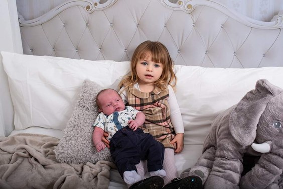 Little girl sitting on a grey bed holding her baby brother