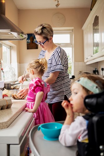 Mum and daughter standing at the kitchen counter cooking with little girl in foreground in a wheelchair