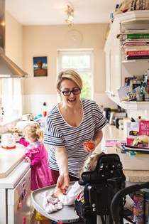 Mum standing in a kitchen passing her young daughter in a wheelchair something