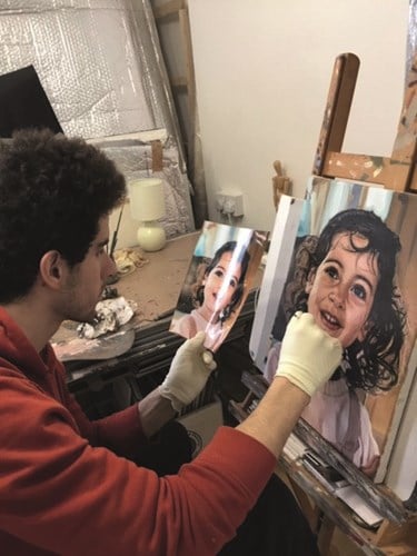 Photo of a young man in a red jumper with dark hair painting a portrait of a young girl with curly brown hair