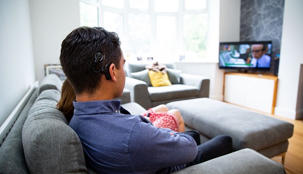 Photo taken over the shoulder over a young man wearing a blue shirt and a cochlear implant. He sits on the sofa beside a woman wearing a red skirt, watching TV with subtitles on.