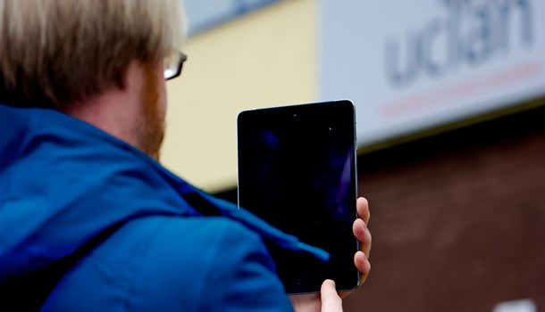 Photo taken over the shoulder of a young white man with blonde hair. The man is outside, holding up a computer tablet.