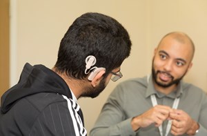 Photo taken over the shoulder of a young man wearing a black striped hoodie, glasses and a cochlear implant. He is watching a man in a grey shirt who is signing to him. The signing man wears a lanyard.