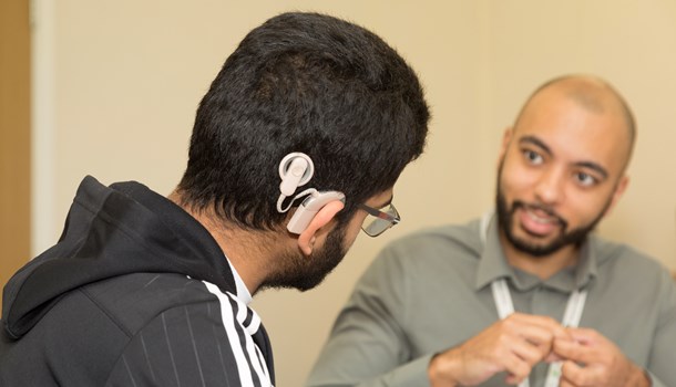 Photo taken over the shoulder of a young man wearing a black striped hoodie, glasses and a cochlear implant. He is watching a man in a grey shirt who is signing to him. The signing man wears a lanyard.