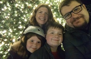 Photo of mother, father, daughter and son standing in front of a tree with fairy lights on. Daughter is wearing a silver cycling helmet
