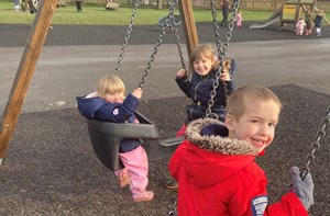 Photo shows three children alongside each other on a set of swings outside. Picture shows two girls in dark blue jackets and a boy, closest to the camera, in a bright red jacket