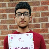Photo of a teenage boy wearing a red jumper standing against a brick wall holding a certificate