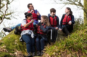 A group of deaf young people wearing life jackets sit on a grassy slope, chatting in sign language.