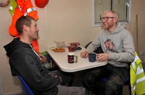 Two construction workers sit at a table laughing. There are mugs of tea, hard hats and high-vis jackets on the table.