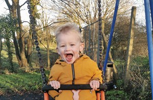 Photo shows a young boy in a yellow jacket and blue trousers swinging on a swing